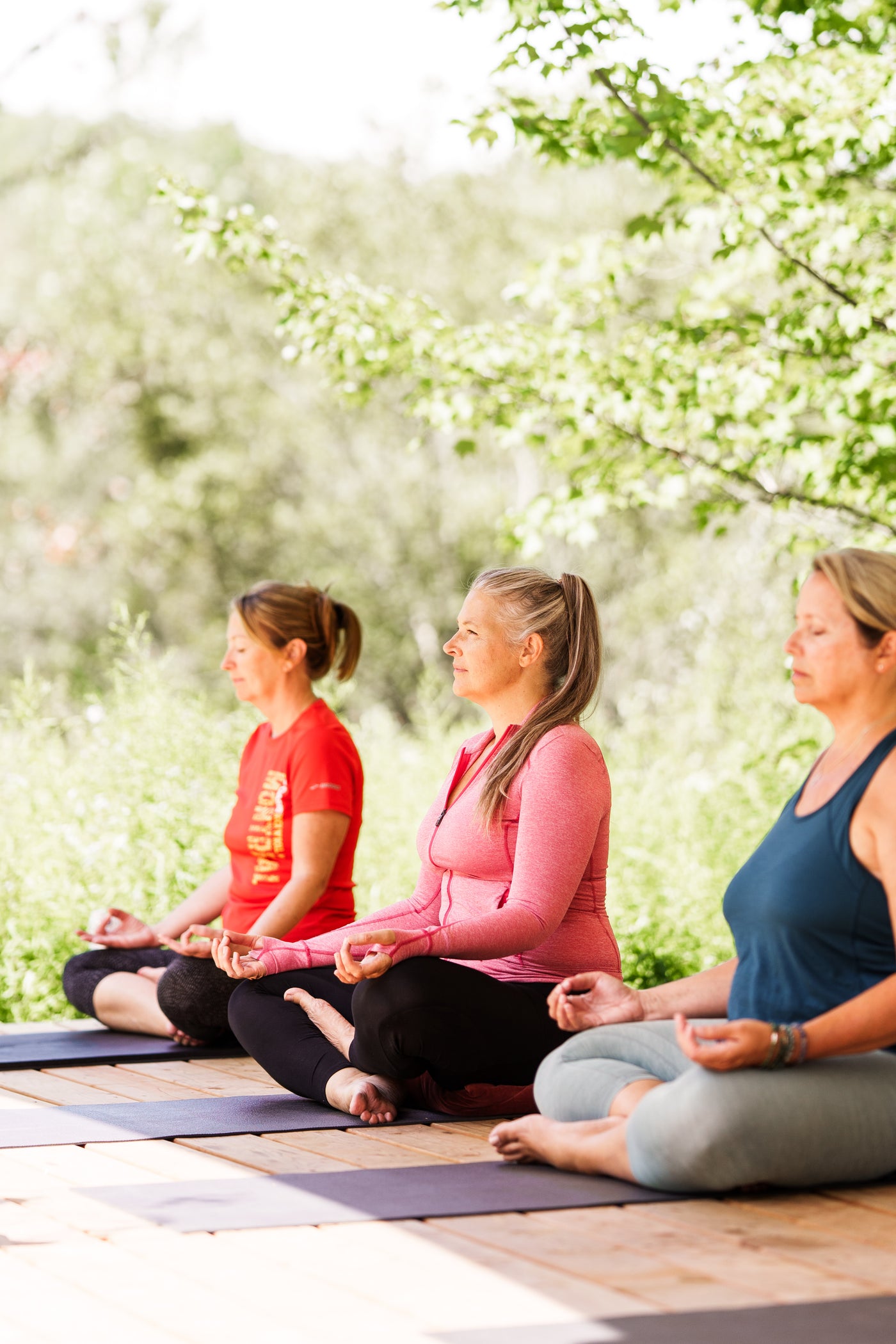 Trois femmes assises en tailleur sur des tapis de yoga, pratiquant la méditation en plein air. Elles sont concentrées, les yeux fermés ou légèrement baissés, avec les mains posées sur les genoux en geste de mudra. Chacune porte des vêtements de sport confortables dans des tons rouge, rose et bleu. La scène se déroule sur une terrasse en bois entourée d'une végétation verdoyante, suggérant un cadre paisible et naturel, propice à la relaxation et à la pleine conscience. La lumière naturelle douce illumine la 