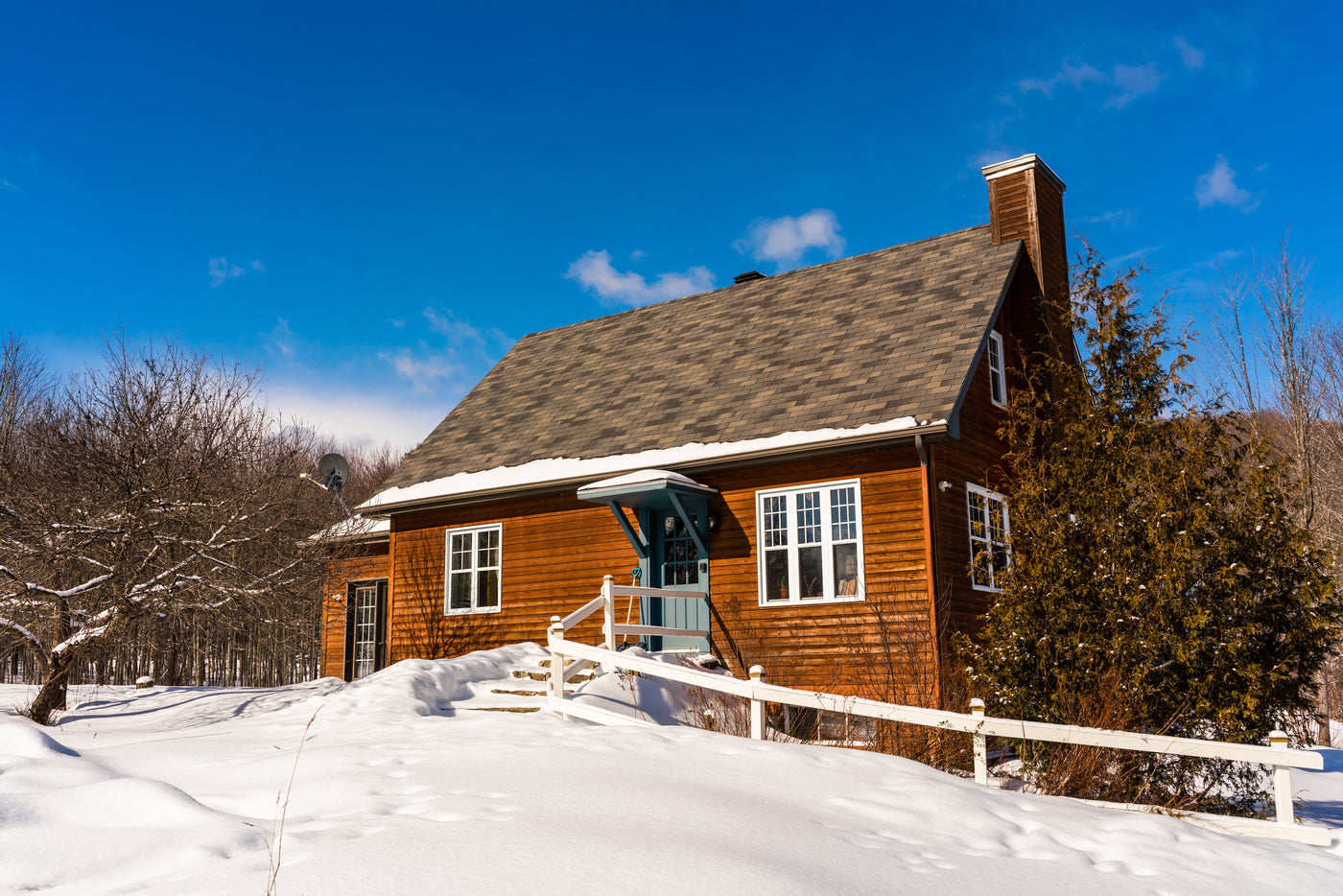 Une maison en bois au style rustique, située dans un paysage hivernal. La toiture est recouverte de bardeaux gris, tandis que le terrain environnant est couvert d'une épaisse couche de neige. La façade de la maison est agrémentée de fenêtres blanches et d'une porte d’entrée bleu pastel, contrastant joliment avec les murs en bois. Un escalier en pierre mène à la porte principale, encadré par une petite barrière blanche. 