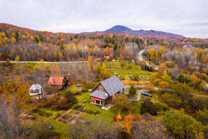 Vue aérienne d’une maison champêtre entourée de paysages automnaux, avec des couleurs vives de feuilles d’automne. En arrière-plan, un dôme géodésique et une grange en bois sont visibles, avec une route sinueuse traversant une forêt colorée. 