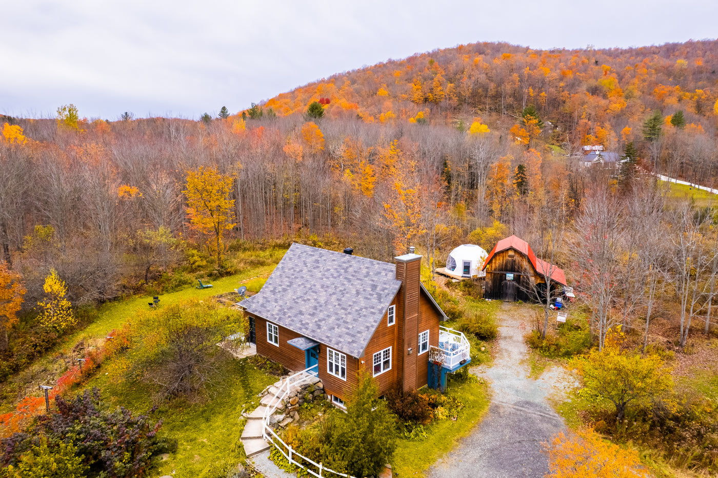 Vue aérienne d'une maison en bois entourée de collines parées de couleurs automnales. Près de la maison, un dôme géodésique blanc et une grange au toit rouge ajoutent au charme rural. La propriété est bordée par une forêt de feuillus aux feuilles jaunes et orange.